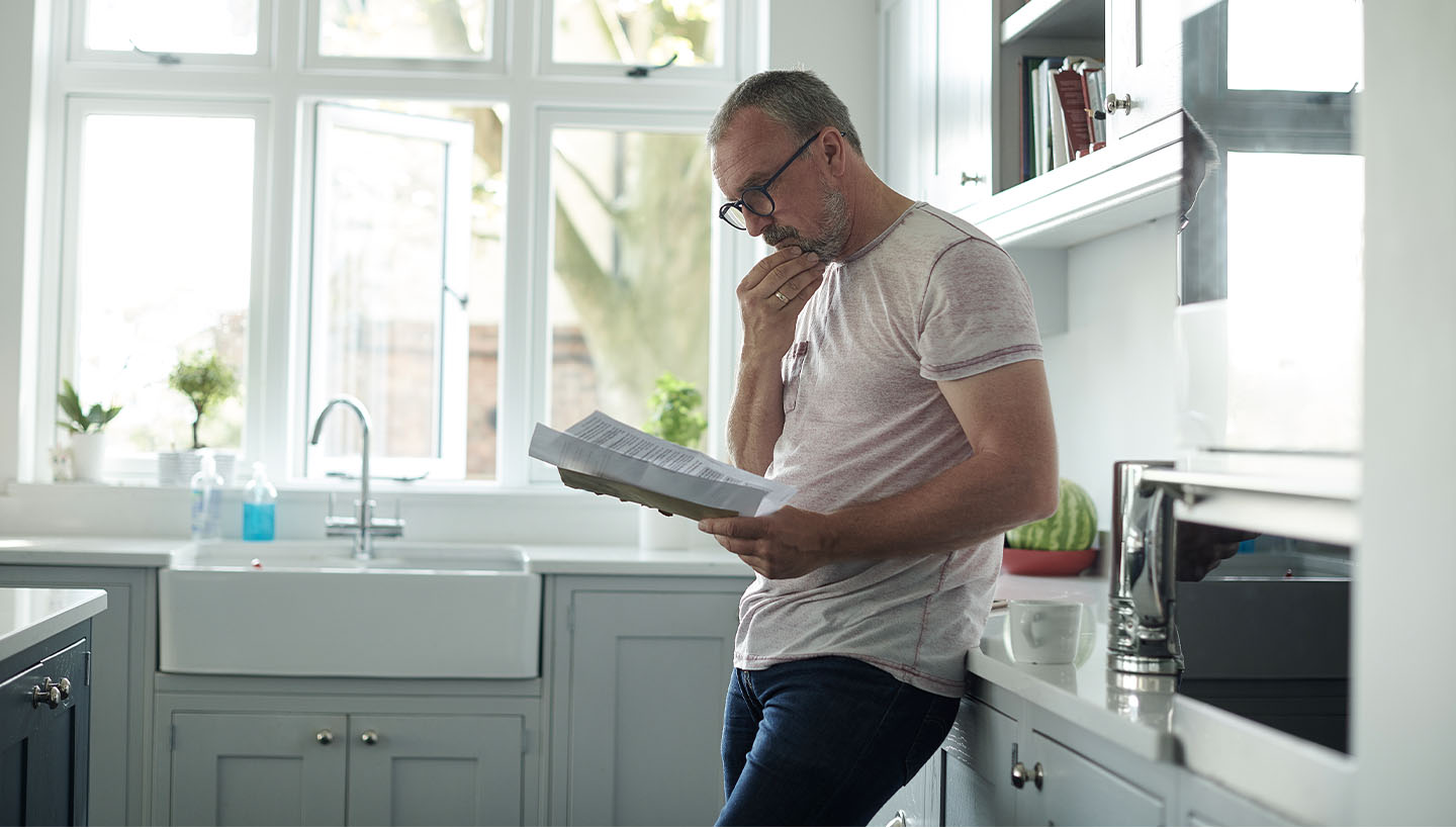 A home inspector standing in a bright kitchen, reading a document thoughtfully while leaning against the counter.