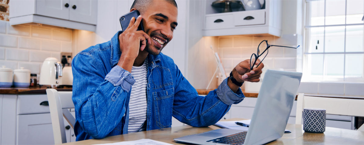 A future insured home inspector sits in a kitchen while holding glasses, talking on the phone, and looking at his laptop–presumably researching affordable insurance for new home inspectors and insurance benefits for AHIT graduates with InspectorPro. 