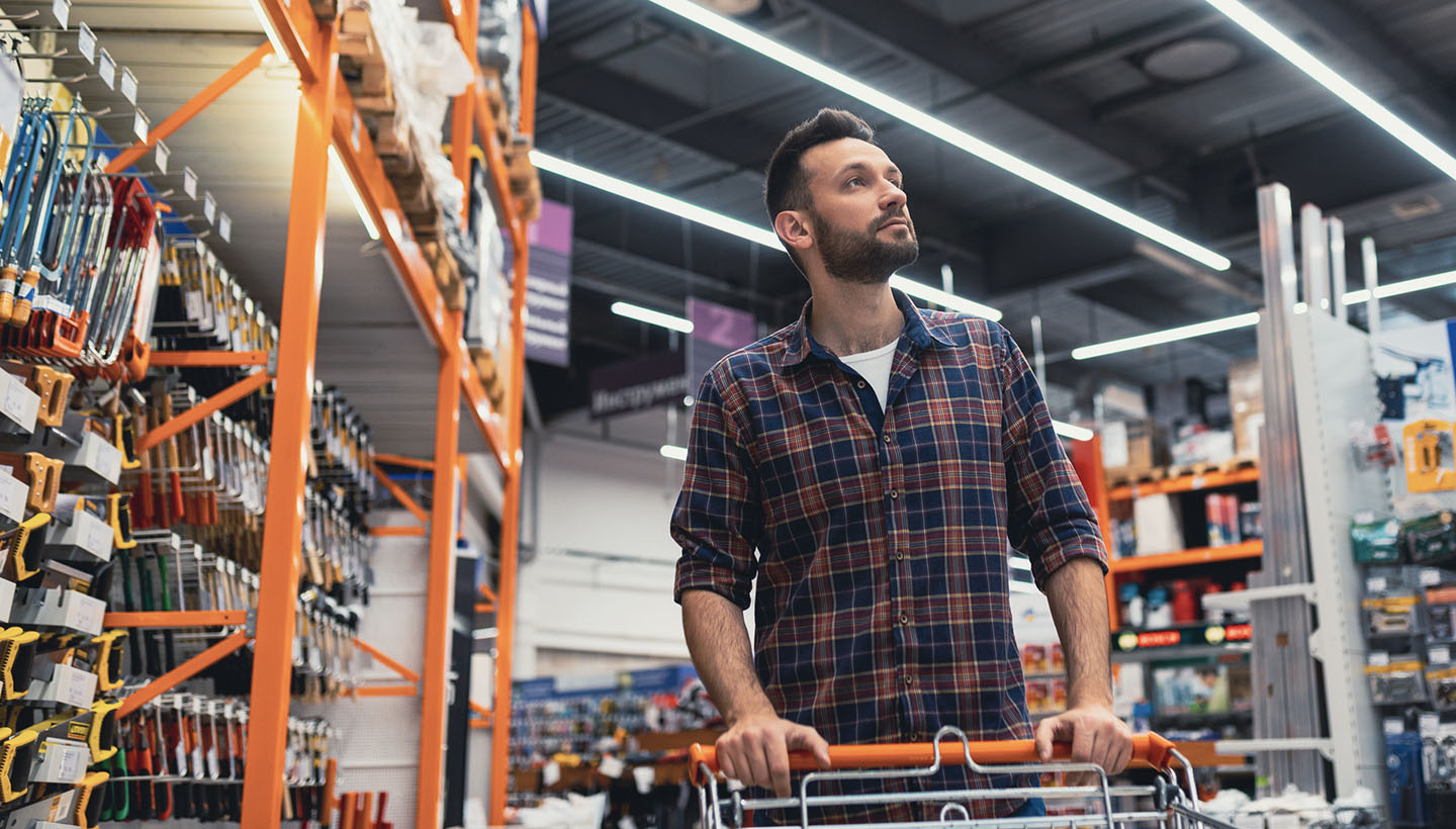 Man in a plaid shirt pushing a shopping cart through a hardware store aisle, surrounded by tools and equipment on shelves