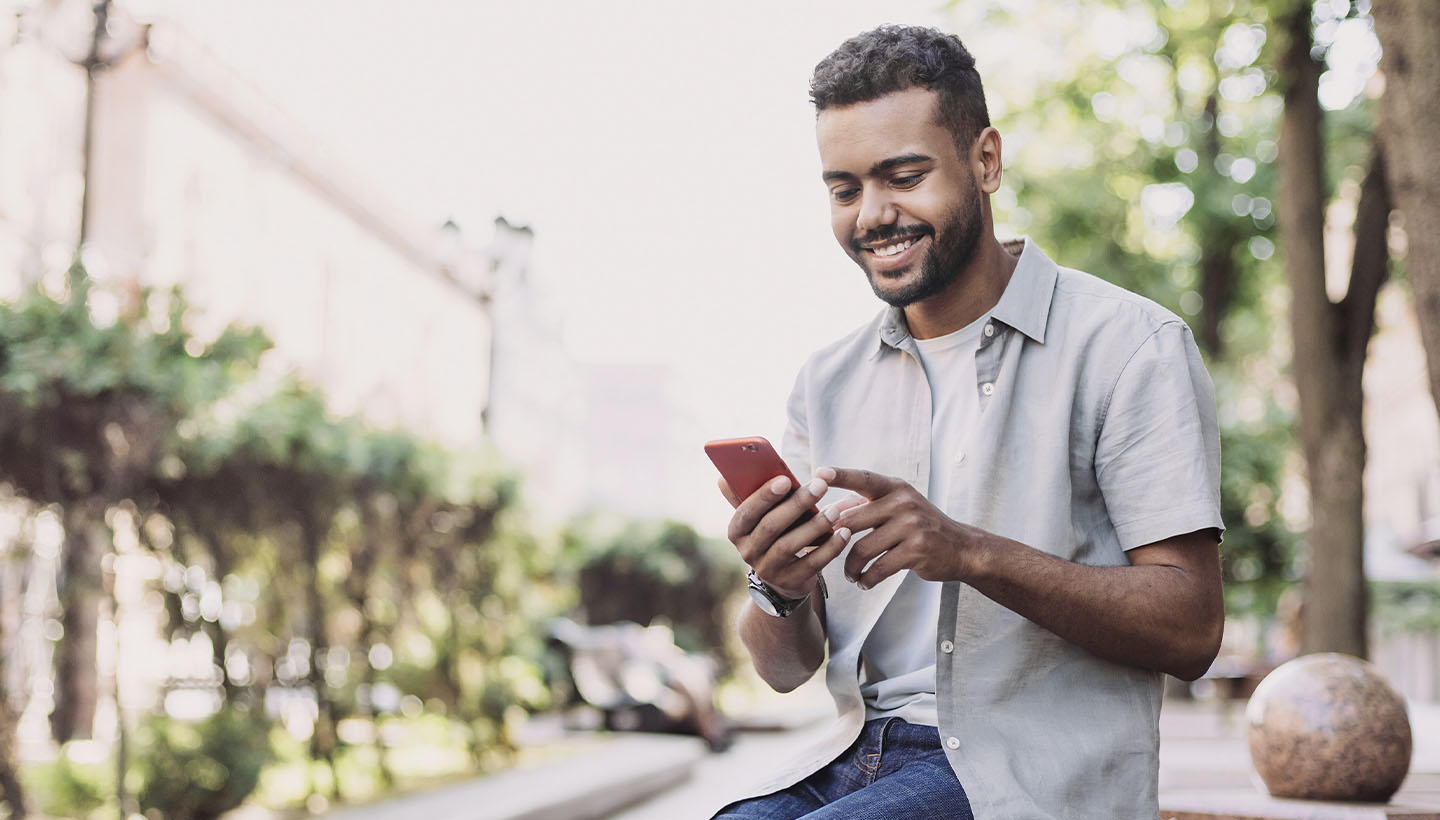1-SocialMediaTipsHomeInspectors-1440×820 Smiling man sitting outdoors, using a smartphone while wearing a light-colored shirt and jeans in a park-like urban setting